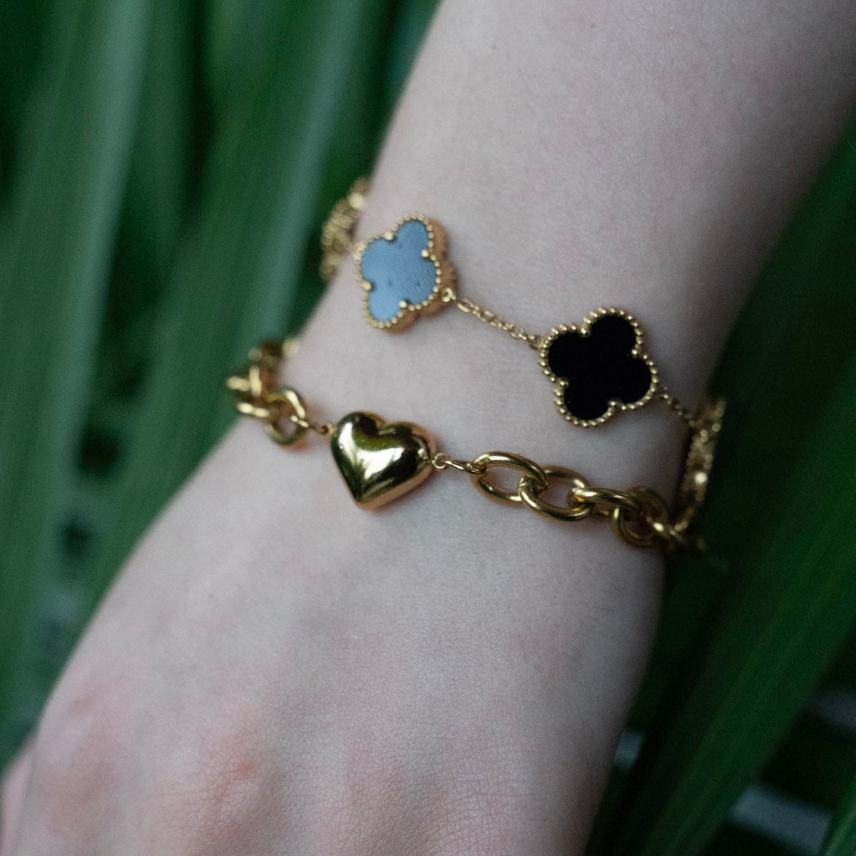 Gold bracelet with heart and clover charms on a wrist against a green leafy background