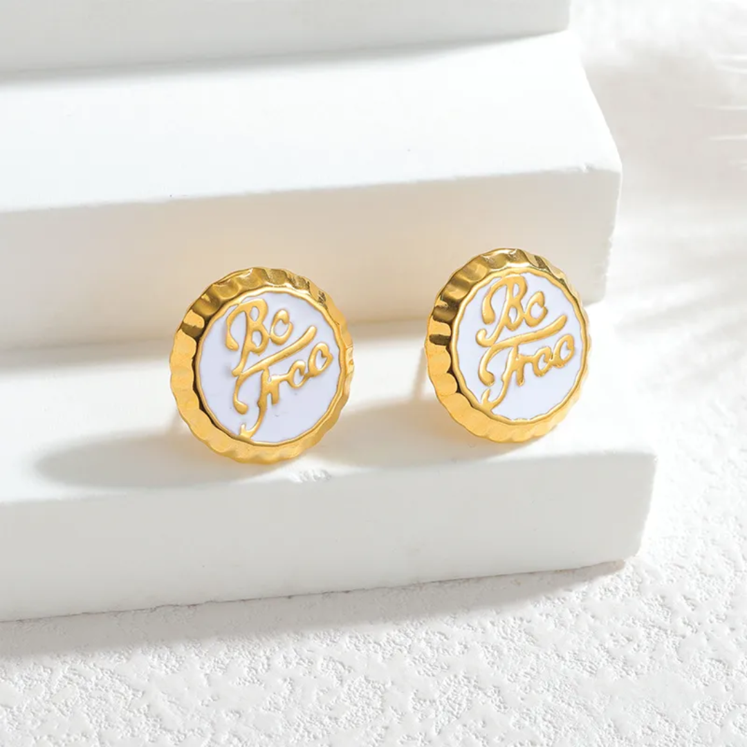 Gold and white earrings with 'Be Free' inscription on a white surface
