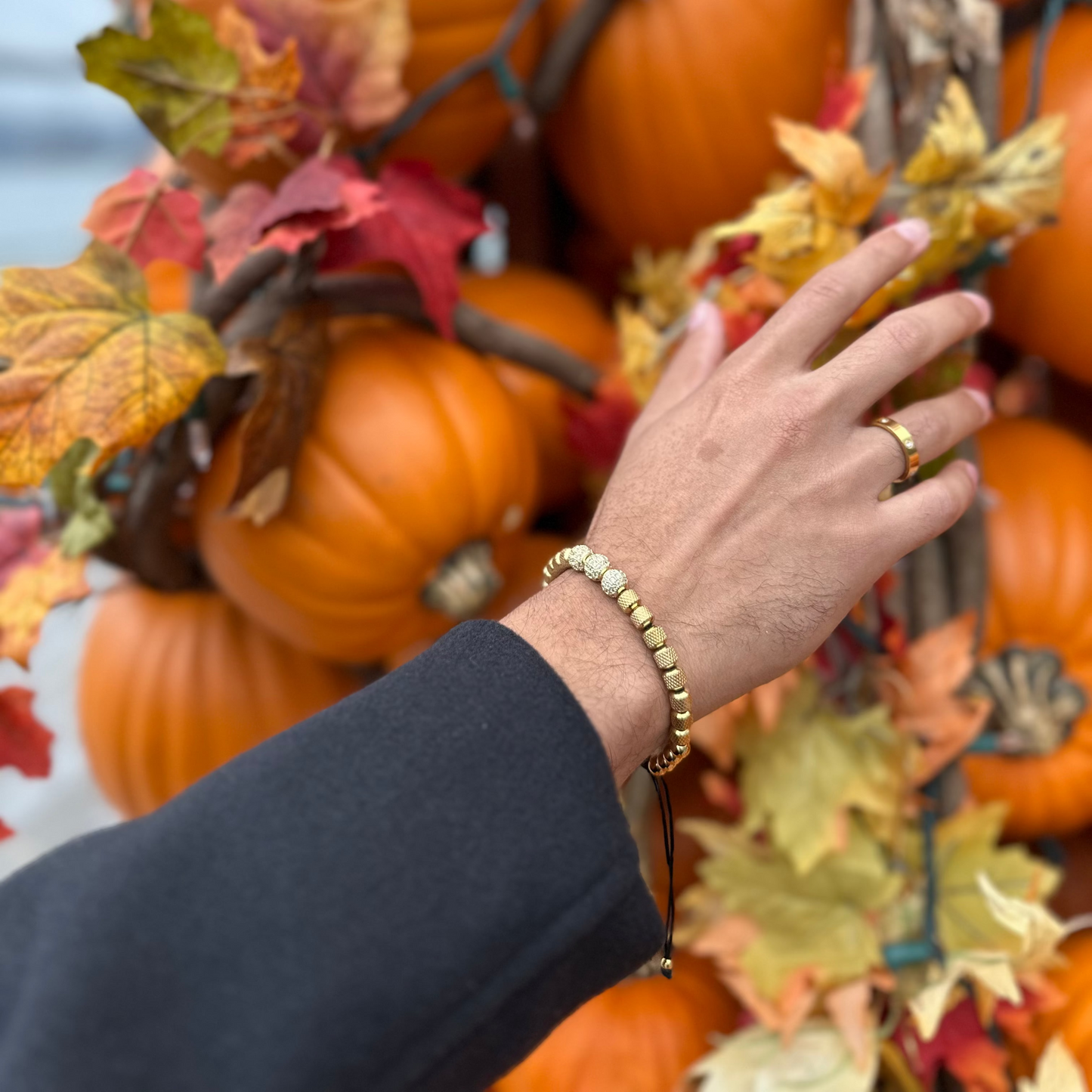 Hand with a gold bracelet reaching towards pumpkins and autumn leaves.