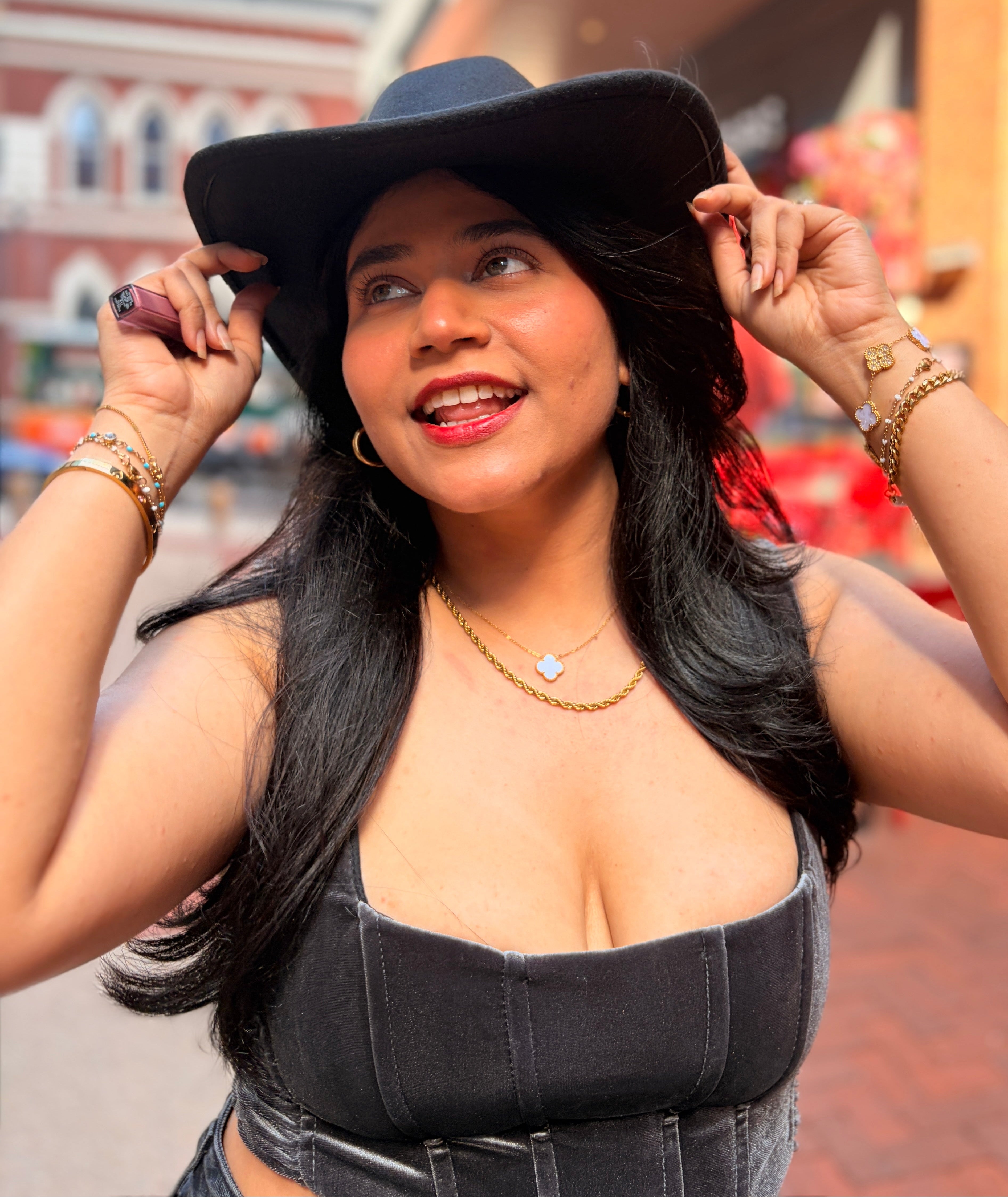 A woman wearing a black wide-brim fedora hat and a black strapless denim corset top, posing outdoors on a brick walkway with a blurred storefront background.
