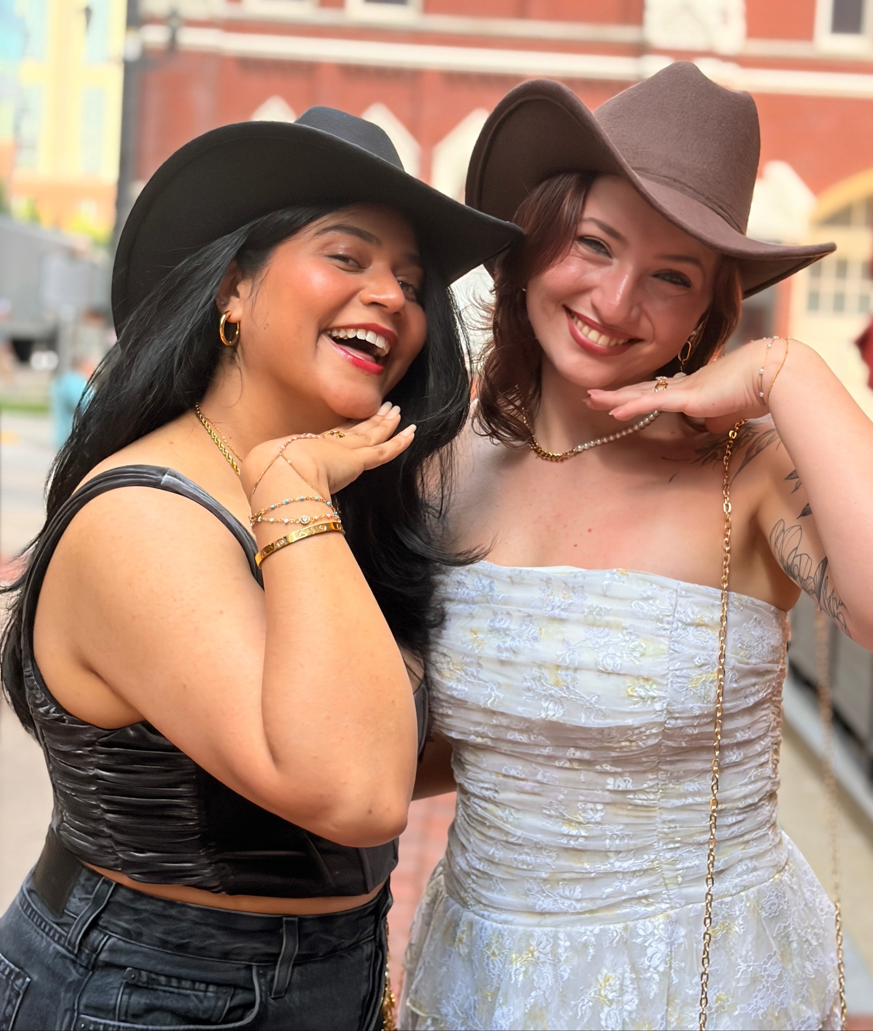 Two women posing outdoors in front of a brick building. The woman on the left wears a black cowboy hat, a black sleeveless top, and dark jeans.