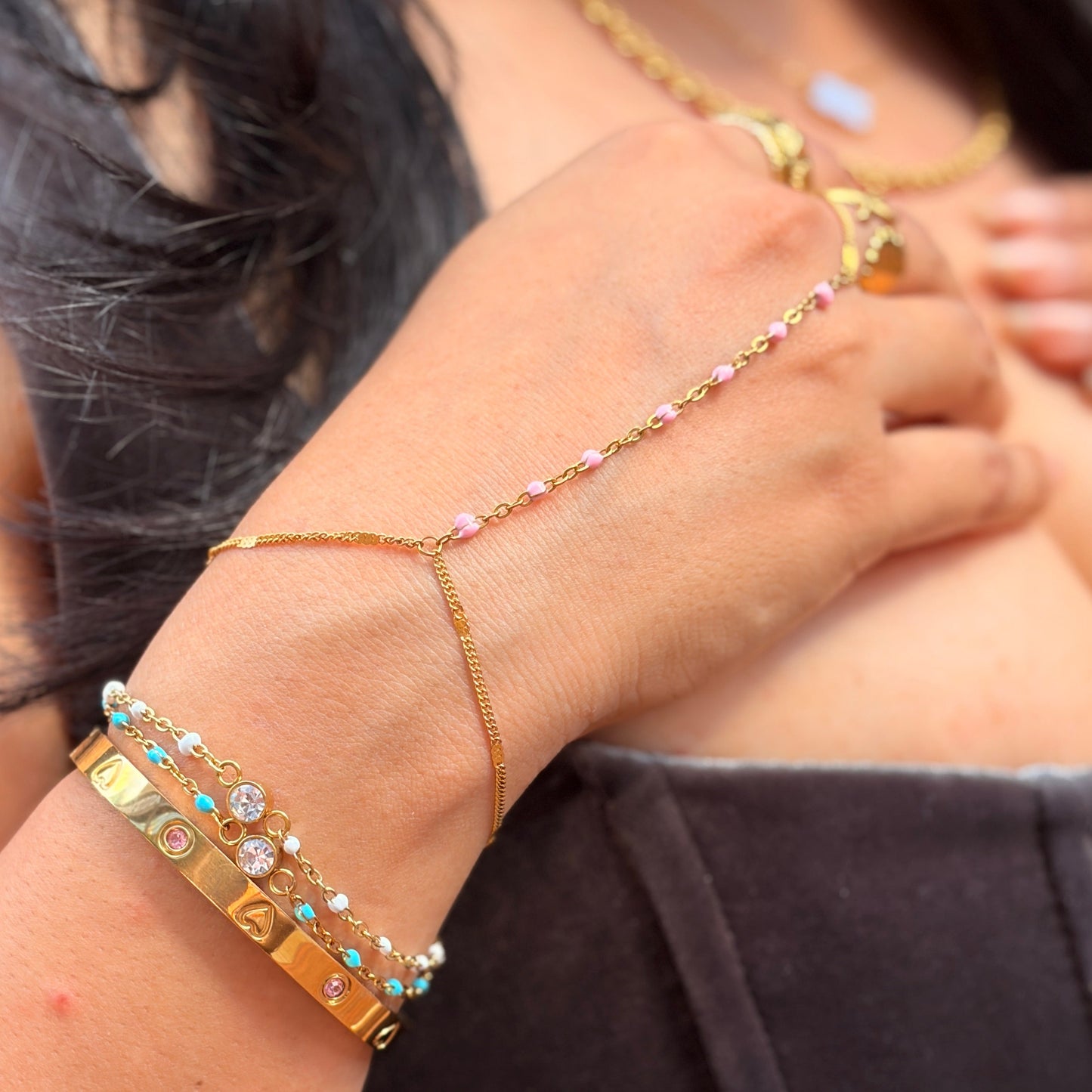 A close-up of a person's wrist and hand wearing a delicate gold chain hand bracelet with small pink and clear rhinestones, paired with stacked gold bracelets on the other wrist.