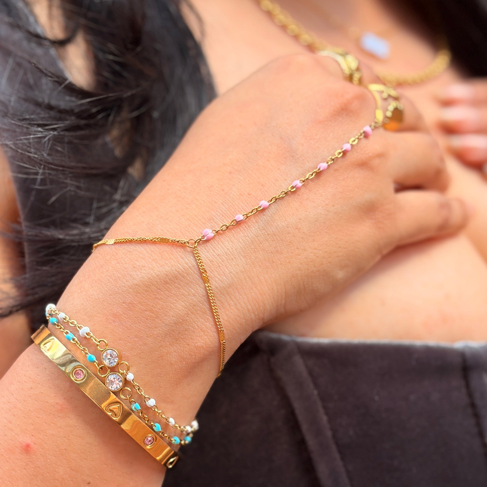 A close-up of a person's wrist and hand wearing a delicate gold chain hand bracelet with small pink and clear rhinestones, paired with stacked gold bracelets on the other wrist.