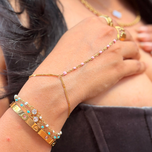 A close-up of a person's wrist and hand wearing a delicate gold chain hand bracelet with small pink and clear rhinestones, paired with stacked gold bracelets on the other wrist.