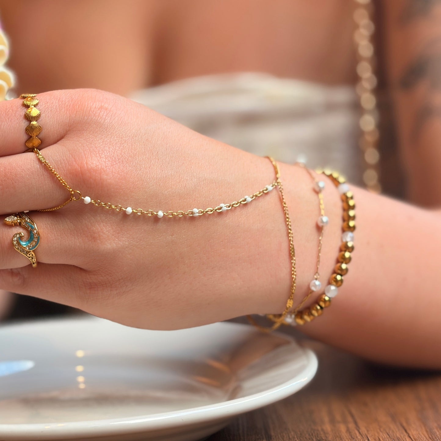 A close-up lifestyle shot of a person holding a frosted cupcake while wearing a gold chain hand bracelet, matching gold ring, and pearl bead bracelets on their wrist, set against a warm cafe background.