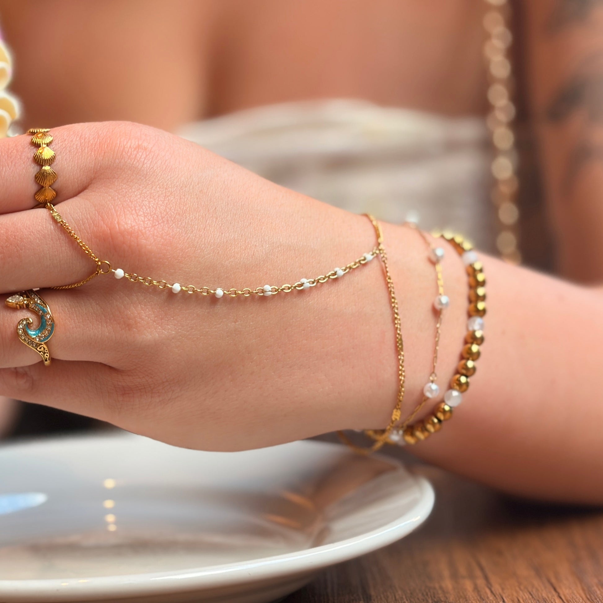 A close-up lifestyle shot of a person holding a frosted cupcake while wearing a gold chain hand bracelet, matching gold ring, and pearl bead bracelets on their wrist, set against a warm cafe background.