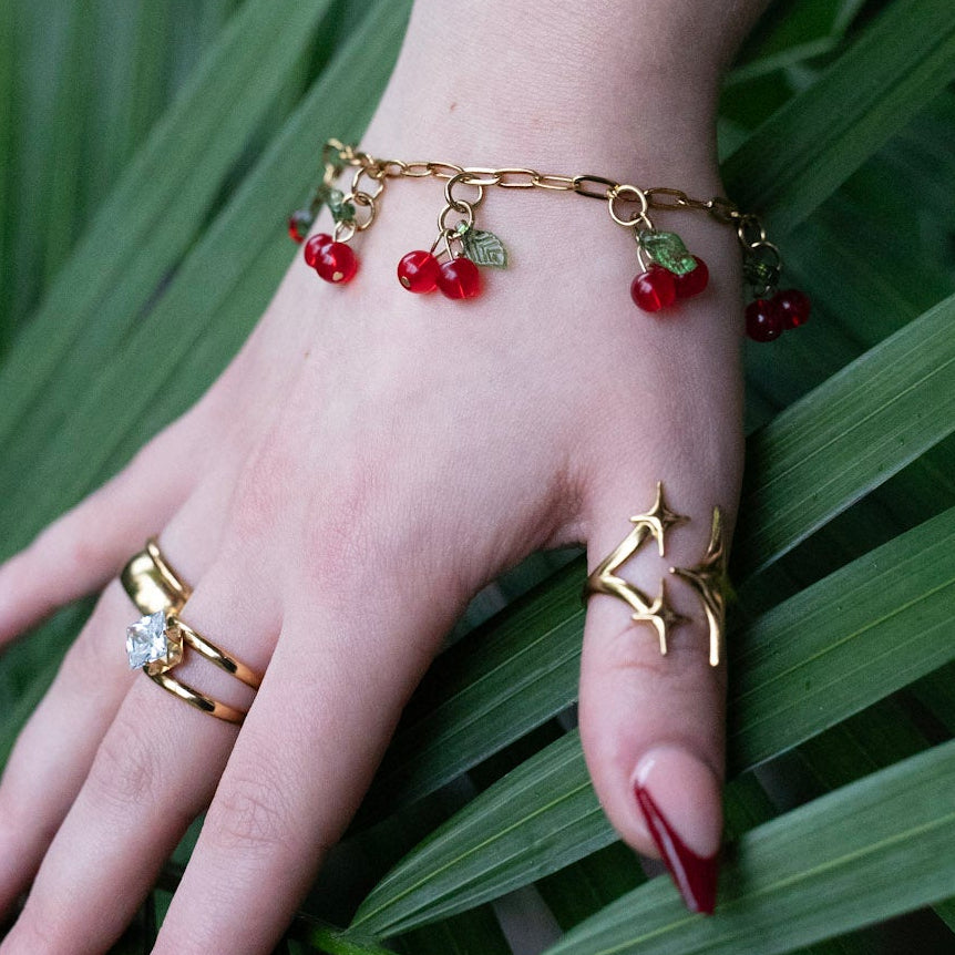 Hand wearing gold rings and a bracelet with red stones on a leafy green background