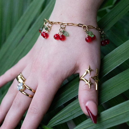 Hand wearing gold rings and a bracelet with red stones on a leafy green background