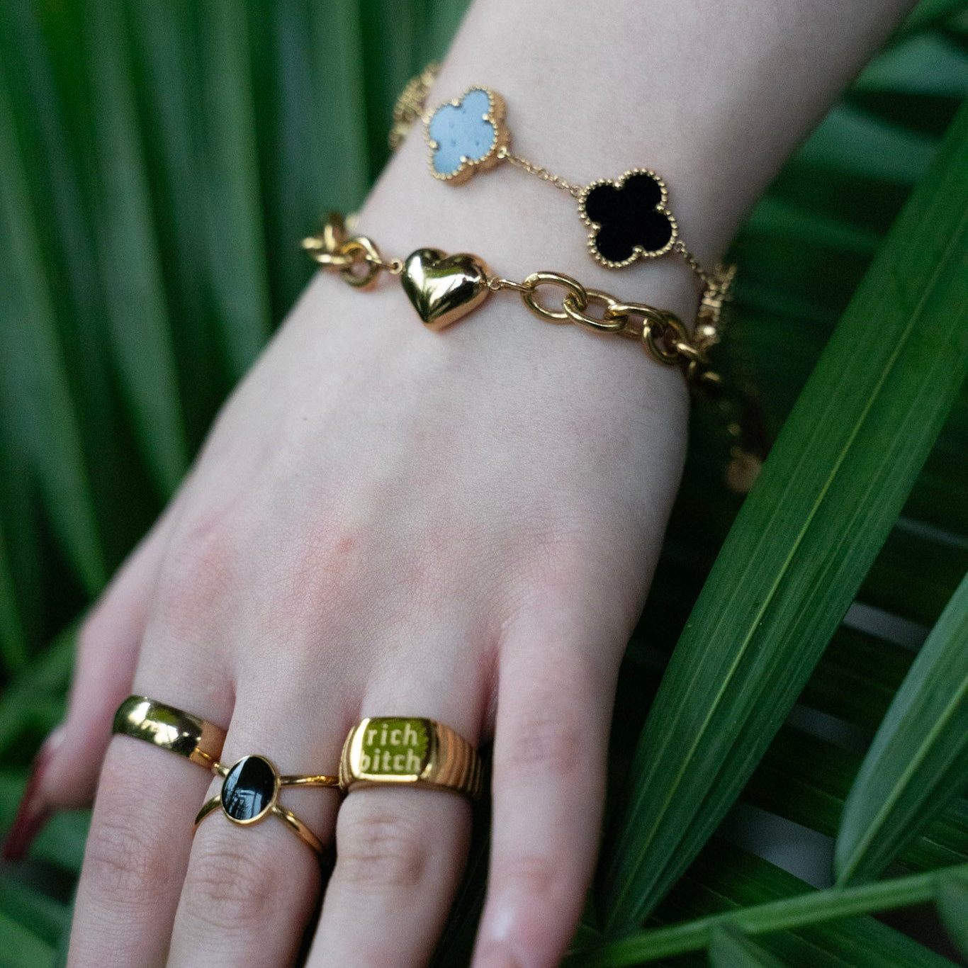 Hand wearing gold jewelry with rings and bracelets on a green leaf background