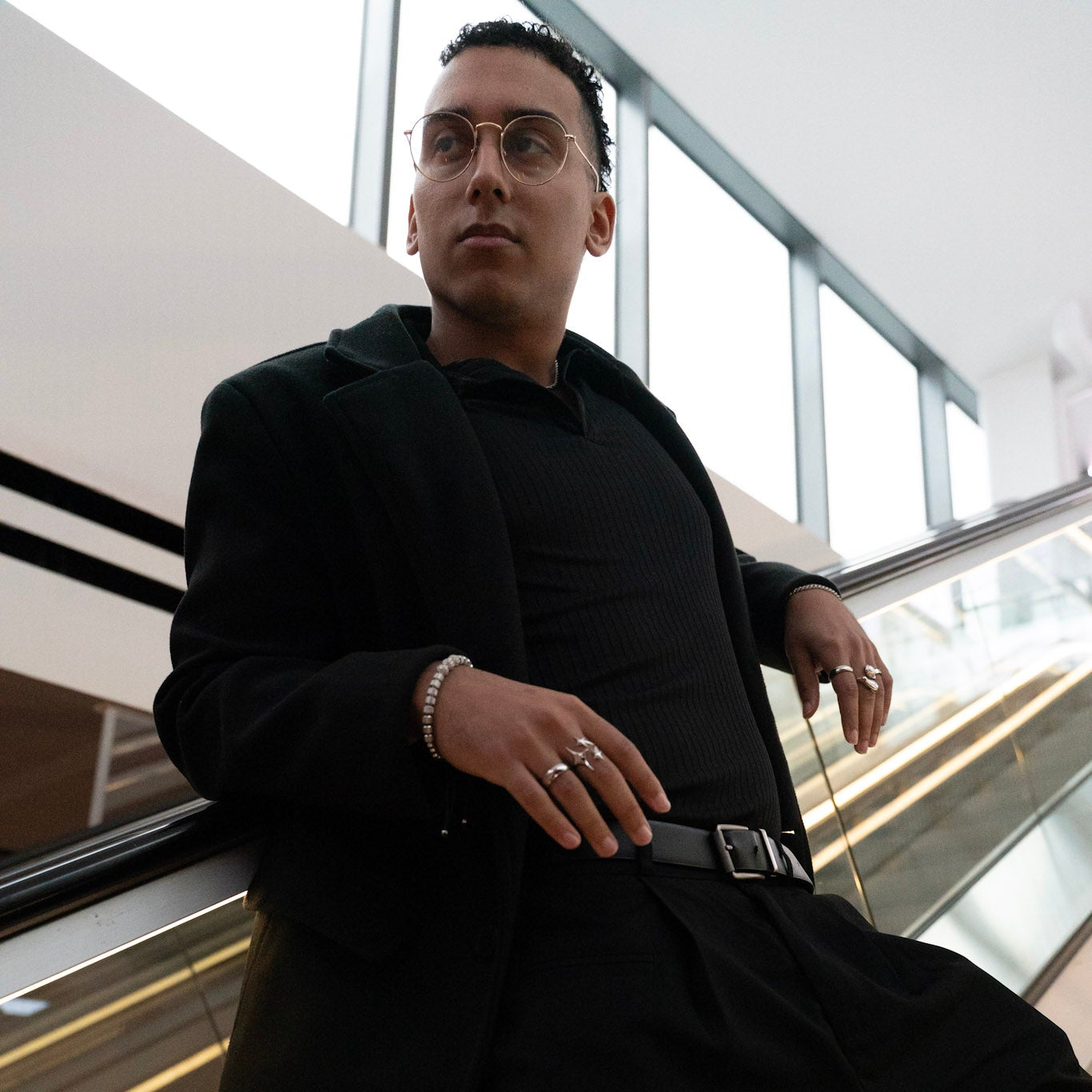 Man in a black coat standing in an indoor setting with large windows.