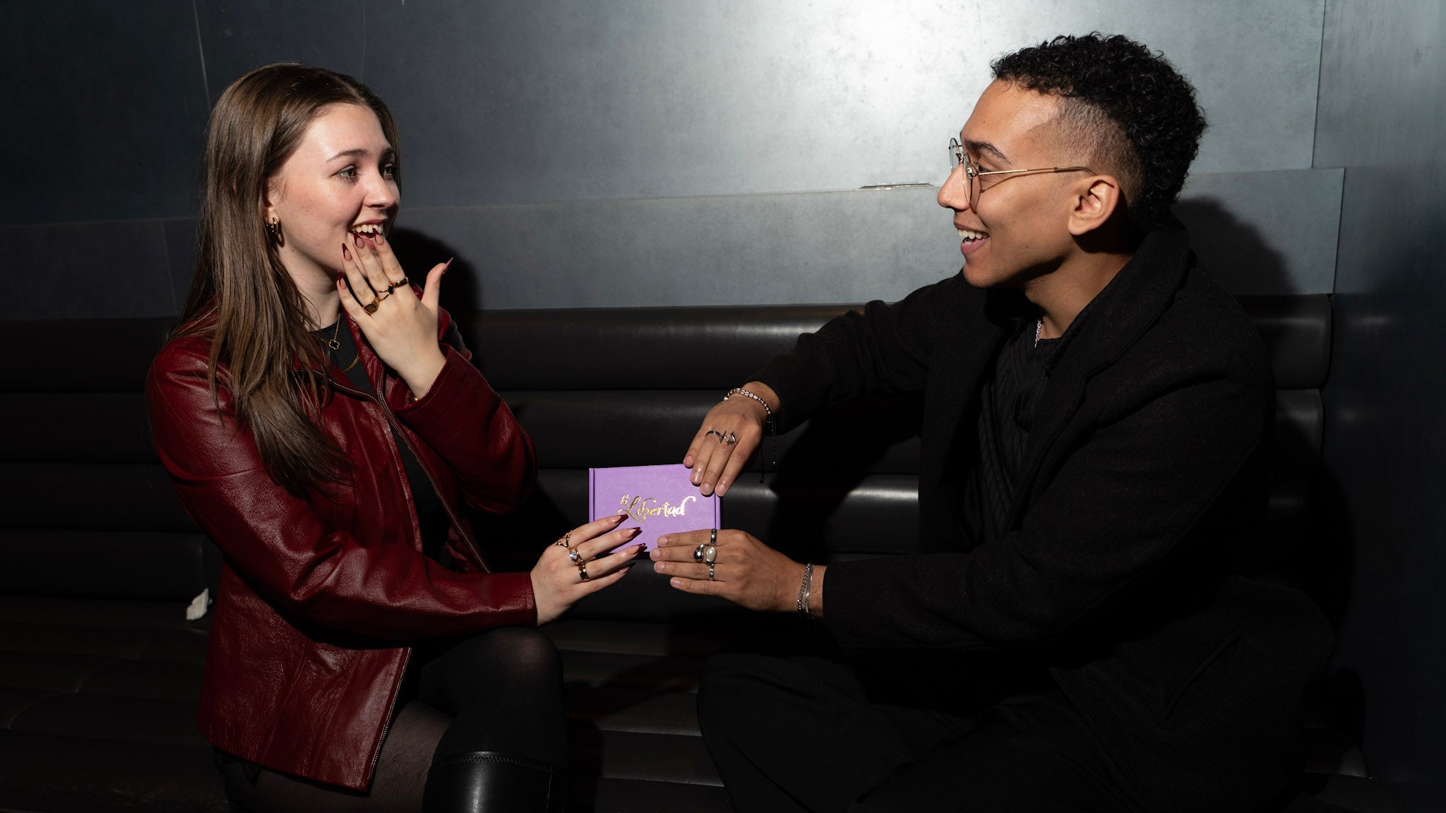 Two people sitting on a bench, one holding a purple box, in a dimly lit setting.